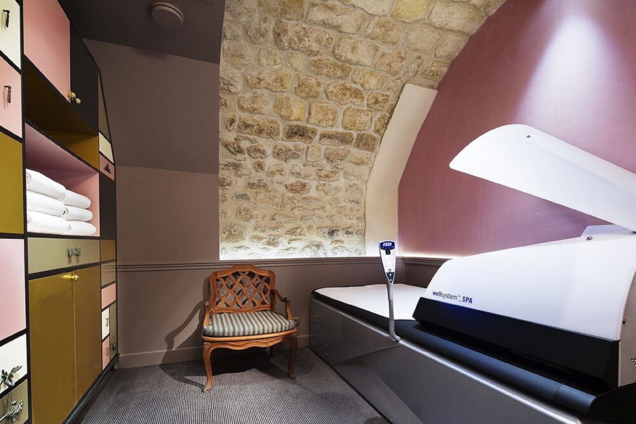 Room with a massage bed on the right hand side, with an arched cover up on the head section for maximum privacy. A chair is next to the bed, and some storage space with towels are on the other side of the room