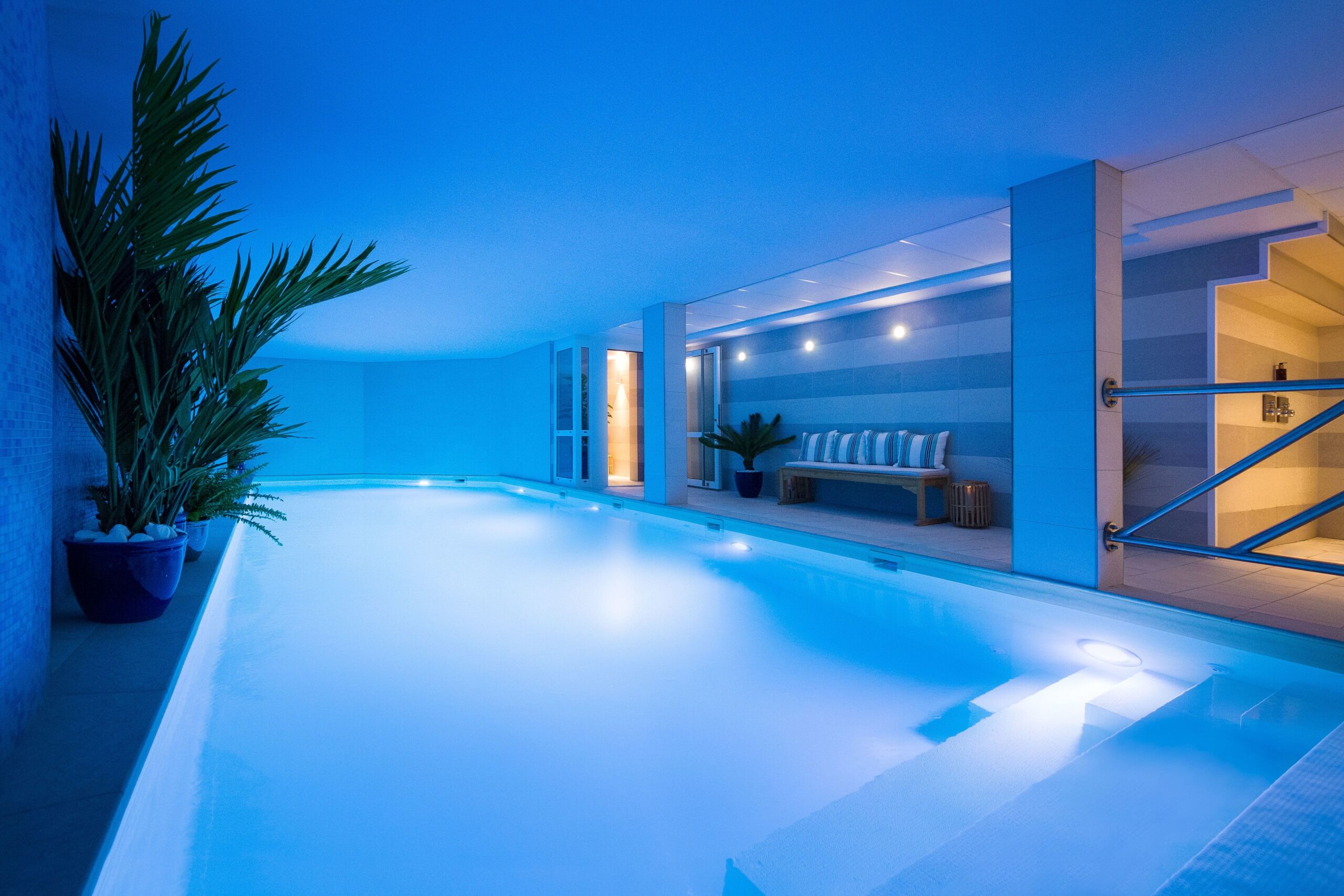 Long rectangular pool, with stairs on one corner. Some plants are placed on one side of the pool and a sofa as well as a couple doors are located on the other side