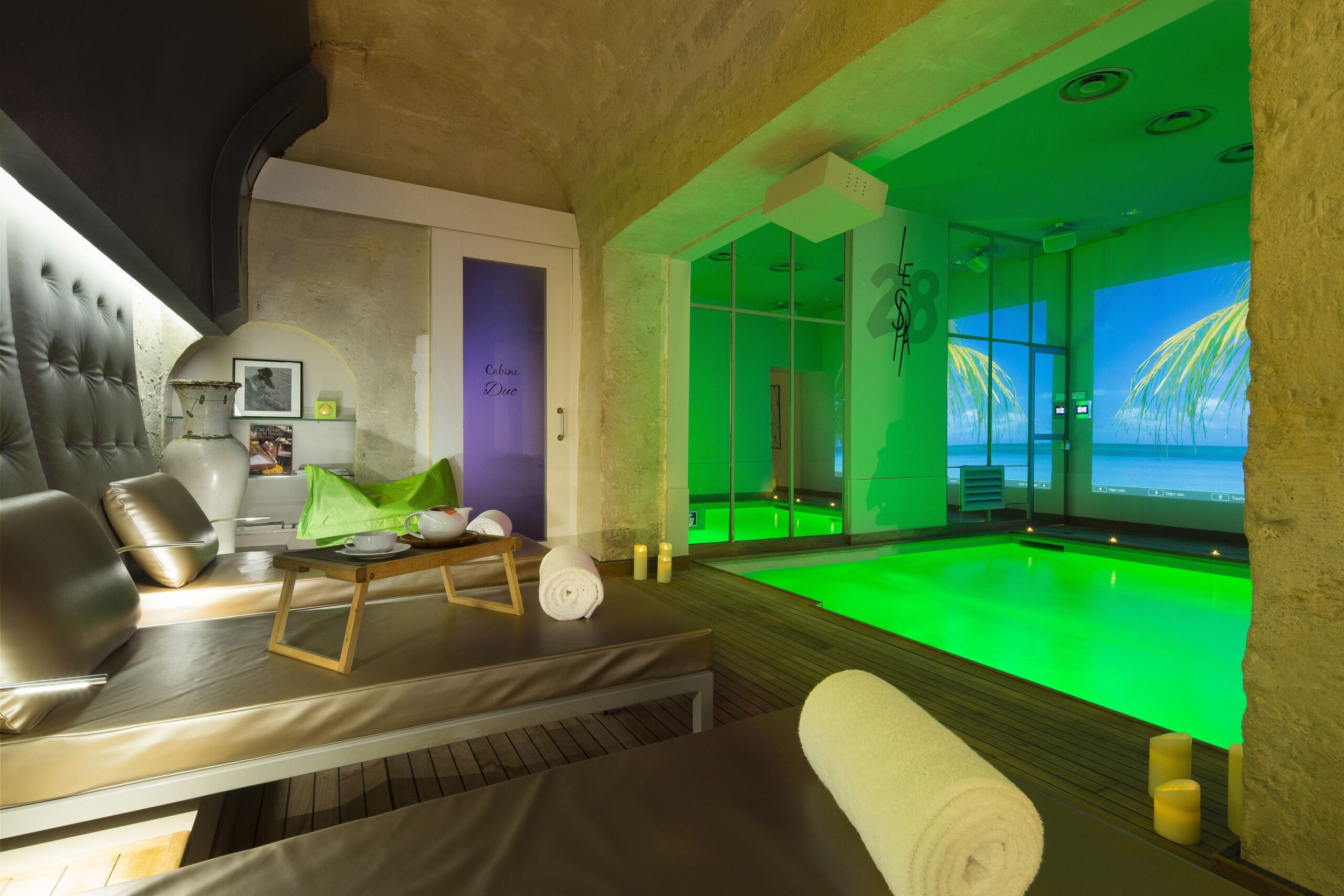 Half of the room is a seating and laying down area with comfortable backrest, and the other side of the room there is a rectangular pool lit up by green lights and a projector can be seen on the furthest wall.