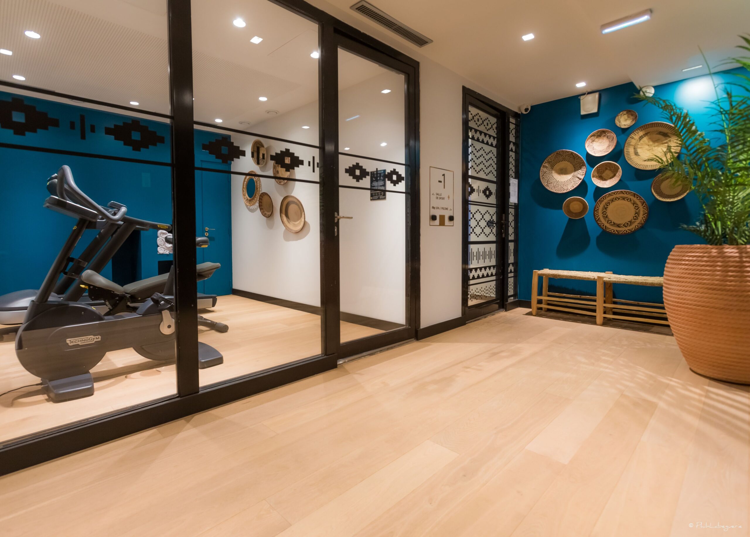 Hallway with a glass door opening up to a Fitness area containing a running and cycling machine. At the end of the hallway on the left there is another door.