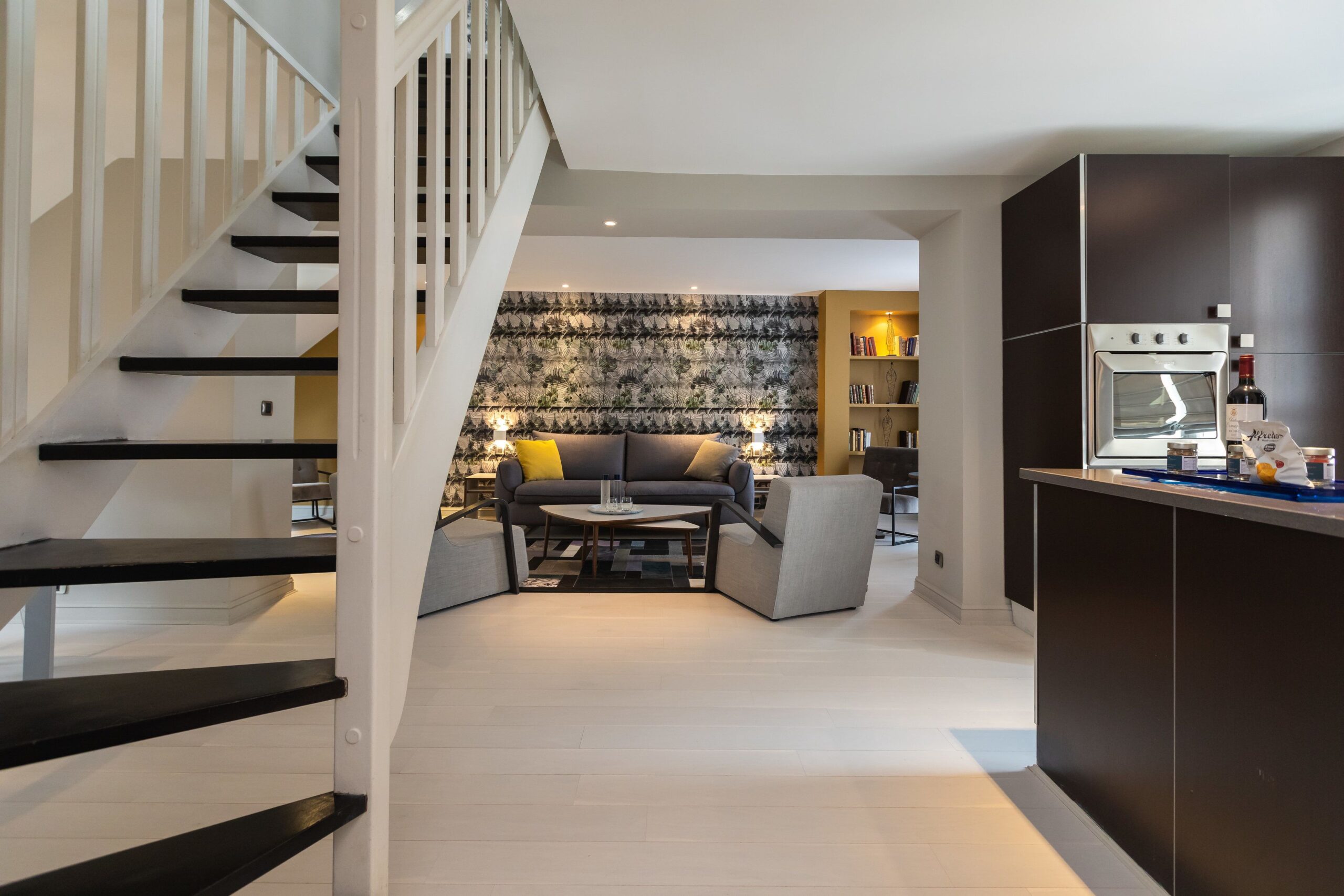 Suite living room where a siting area can be seen at the far end, with a kitchen like area on the right and some stairs going up on the right 