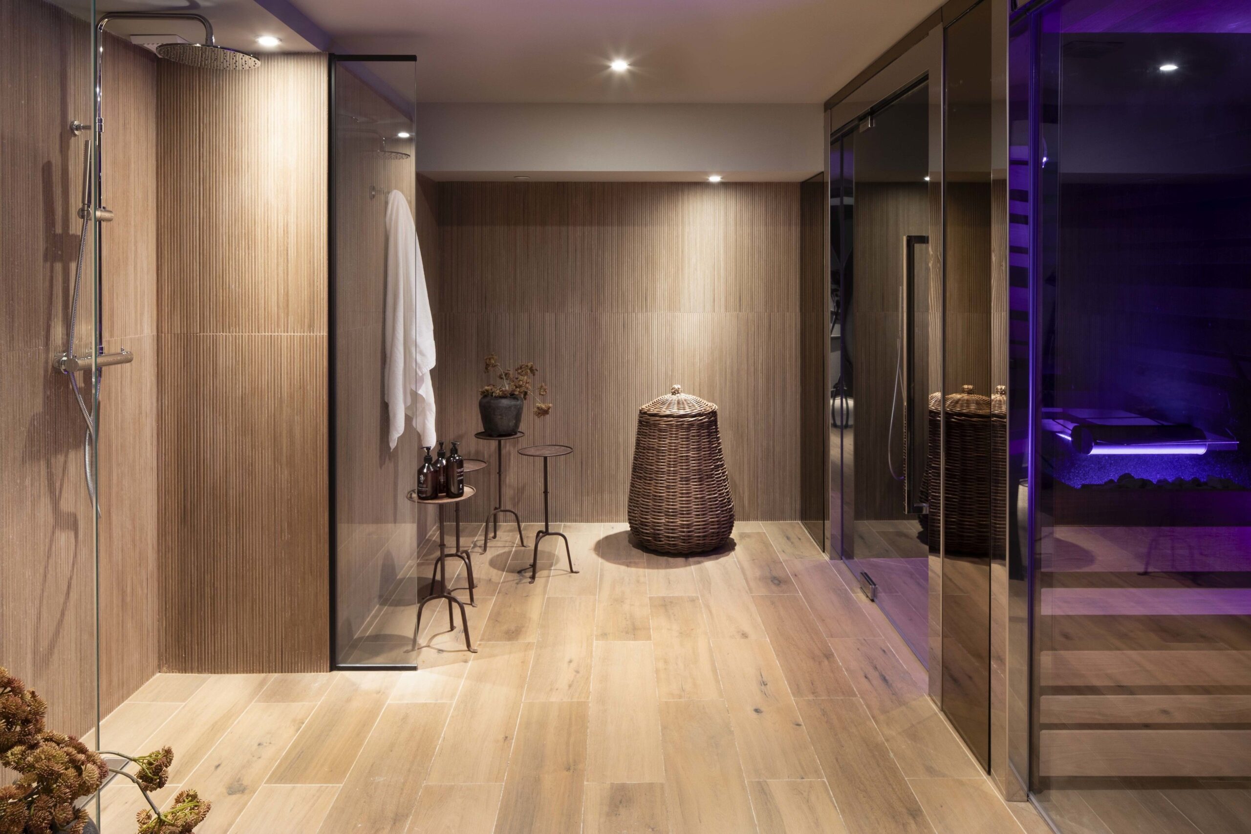 A spa area where a shower can be seen on the left wall, towards the end of the room there are some tables with some decoration and creams as well as a place to hang a towel, and finally on the right hand side can be seen glass doors to what appears to be a hot tub illuminated by purple lights