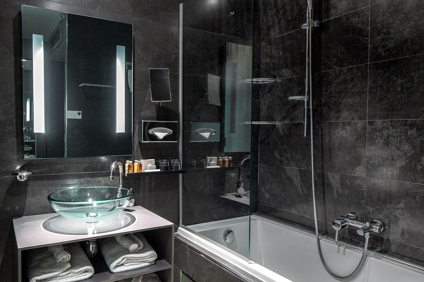 The bathroom features black-colored walls. On the right, there is a tub with a shower head and a glass half-door. Next to it, the sink is positioned underneath a mirror, with towel storage above.