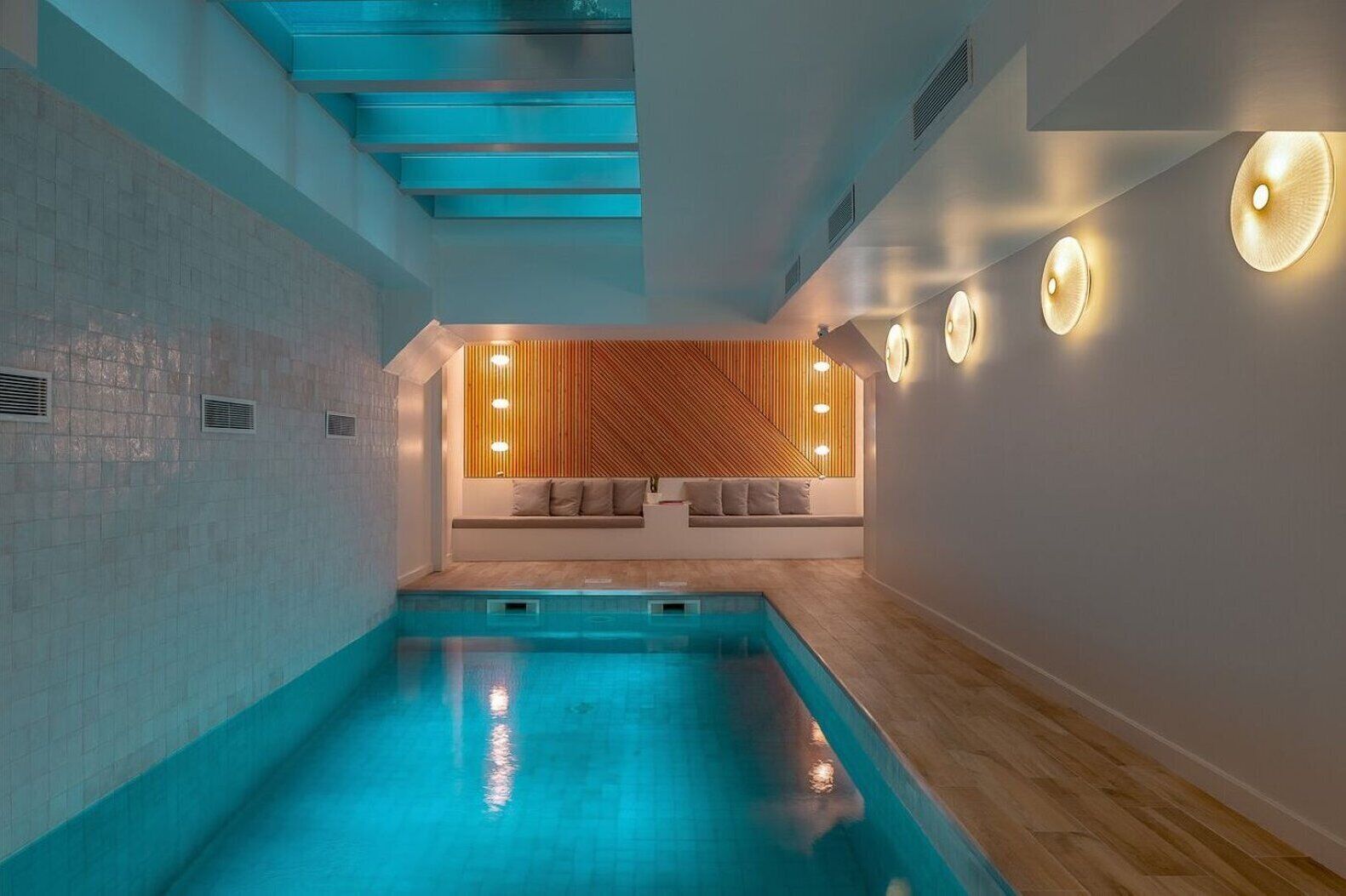 Long rectangular pool with sofas on the far end, a wooden like floor is surrounding the pool on one side, and lights illuminate the way